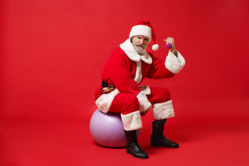 Elderly gray-haired mustache bearded Santa man in Christmas hat posing isolated on red background. Happy New Year 2020 celebration holiday concept. Mock up copy space. Sit on fitball, hold dumbbells.
