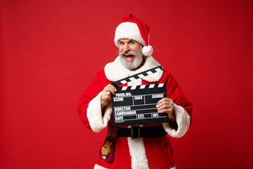 Elderly gray-haired mustache bearded Santa man in Christmas hat posing isolated on red background. New Year 2020 celebration concept. Mock up copy space. Hold classic black film making clapperboard.
