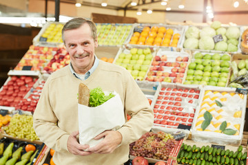 Waist up portrait of smiling senior man holding paper bag with groceries while standing against fruit and vegetable stand at market, copy space