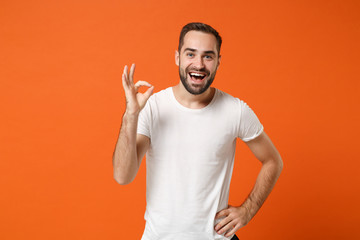 Excited cheerful young man in casual white t-shirt posing isolated on bright orange wall background, studio portrait. People sincere emotions lifestyle concept. Mock up copy space. Showing OK gesture.