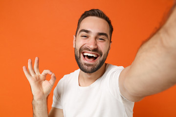 Fototapeta premium Close up of laughing young man in casual white t-shirt posing isolated on orange wall background. People lifestyle concept. Mock up copy space. Doing selfie shot on mobile phone, showing OK gesture.