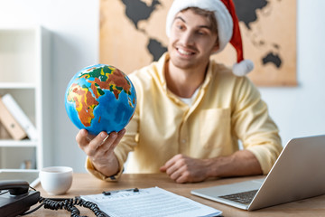 selective focus of cheerful travel agent in santa hat holding globe while sitting at workplace