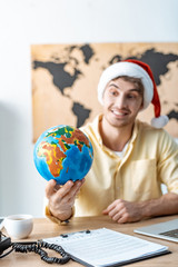 selective focus of smiling travel agent in santa hat holding globe while sitting at workplace