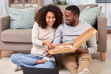 leisure, people and food concept - happy african american couple with laptop computer eating takeaway pizza at home