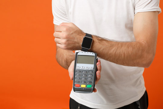 Cropped image of man in white t-shirt posing isolated on orange background. People lifestyle concept. Mock up copy space. Wear smart watch hold bank terminal to process acquire credit card payments.
