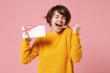 Joyful young brunette woman girl in yellow sweater posing isolated on pastel pink background studio portrait. People lifestyle concept. Mock up copy space. Hold gift certificate, doing winner gesture. - obrazy, fototapety, plakaty