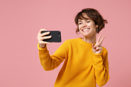 Smiling young brunette woman girl in yellow sweater posing isolated on pastel pink background. People lifestyle concept. Mock up copy space. Doing selfie shot on mobile phone, showing victory sign.