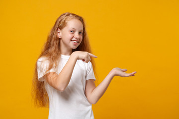 Smiling little ginger kid girl 12-13 years old in white t-shirt isolated on yellow background children portrait. Childhood lifestyle concept. Mock up copy space. Pointing index finger and hand aside.