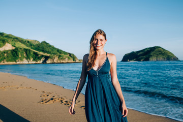 Positive attractive woman in dress walking along sunny seashore