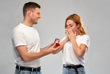 love, couple, proposal and people concept - man giving diamond engagement ring in little red box to happy woman over grey background