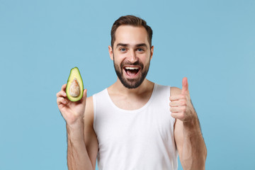 Close up bearded young man 20s years old in white shirt hold half of avocado isolated on blue...
