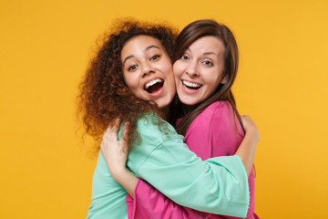 Two cheerful funny women friends european and african american girls in pink green clothes posing isolated on yellow background. People lifestyle concept. Mock up copy space. Hugging, looking camera.