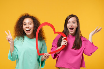 Two women friends european african american girls in pink green clothes posing isolated on yellow...