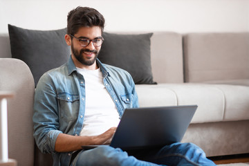 Young happy caucasian hipster with eyeglasses sitting on floor in living room, holding laptop in lap and typing on it.