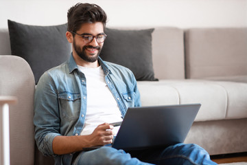 Smiling caucasian hipster sitting on floor in living room, holding laptop in lap and using credit card for online shopping or paying bills.