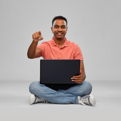 technology, internet, communication and people concept - happy indian man with laptop computer sitting on floor and pointing fnger to camera over grey background