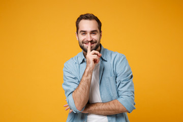 Smiling young bearded man in casual blue shirt posing isolated on yellow orange wall background. People lifestyle concept. Mock up copy space. Saying hush be quiet with finger on lips shhh gesture.