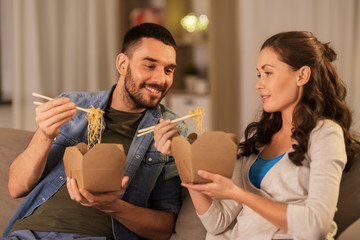 fast food and people concept - happy couple eating takeaway noodles with chopstick at home in evening