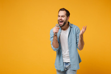 Handsome young man in casual blue shirt posing isolated on yellow orange wall background, studio portrait. People lifestyle concept. Mock up copy space. Sing song in microphone, keeping eyes closed.