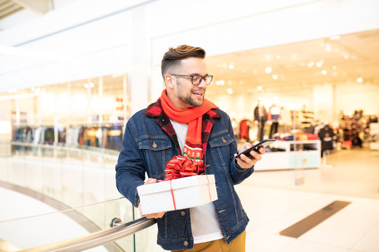 Charming hipster guy smiling and chatting on his smart-phone. He is wearing glasses and a scarf. He is waiting for a friend while he is holding a Christmas gift. He is standing in the shopping mall.