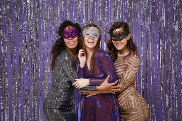 Beautiful mysterious women in fancy sparkling dresses carnival masks posing isolated over vibrant purple violet silver background. People spending time on happy birthday new year holiday party disco.