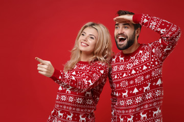Funny young couple guy girl in Christmas sweaters posing isolated on red background studio portrait. Happy New Year 2020 celebration party concept. Mock up copy space. Pointing index fingers aside.