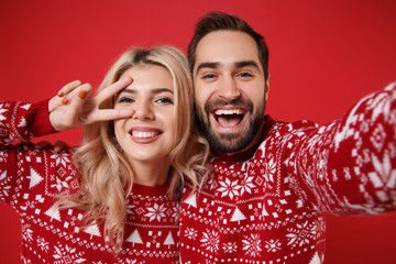 Close up of merry young couple guy girl in Christmas sweaters posing isolated on red background. Happy New Year 2020 celebration party concept. Mock up copy space. Doing selfie shot on mobile phone.