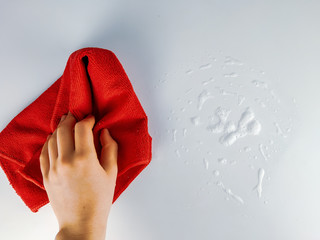 Top view of a female hand holding a red wipe rag and cleaning the bubbly foam on a white surface made from detergent or soap
