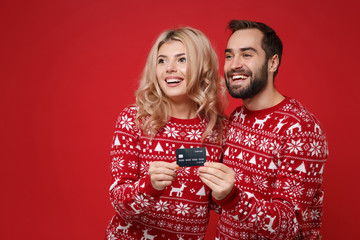 Smiling young couple guy girl in Christmas knitted sweaters posing isolated on bright red background. Happy New Year 2020 celebration holiday party concept. Mock up copy space. Hold credit bank card.