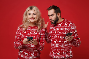 Shocked smiling young couple guy girl in Christmas sweaters posing isolated on red background. New Year 2020 celebration holiday concept. Mock up copy space. Using mobile phone typing sms message.