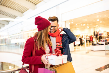 Happy young caucasian couple standing in the shopping mall. Girlfriend is holding a Christmas gift and carrying bags. Boyfriend is looking at her smiling.