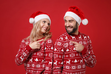 Pretty young couple guy girl in Christmas sweaters, Santa hat posing isolated on red background. New Year 2020 celebration holiday party concept. Mock up copy space. Point index fingers at each other.
