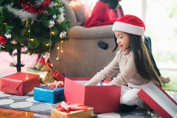 Young Asian family celebration in Christmas day, daughter siting on floor which open give box which smiling felling happy, parent sitting on sofa in living room at home. Merry Xmas happy new year.