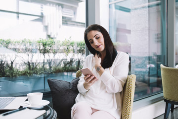 Pleasant woman watching at smartphone sitting in chair with pillow at table