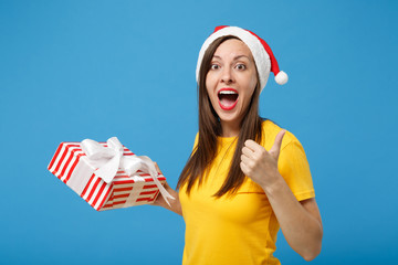 Excited woman Santa girl in Christmas hat posing isolated on blue background. New Year 2020 celebration holiday concept. Mock up copy space. Holding red striped present box with gift showing thumb up.