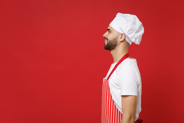 Side view of smiling young bearded male chef cook or baker man in striped apron white t-shirt toque chefs hat posing isolated on red background in studio. Cooking food concept. Mock up copy space.