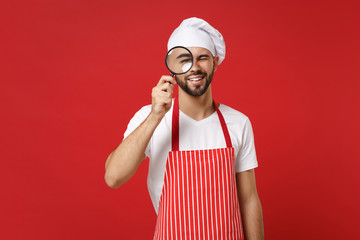Smiling young bearded male chef cook or baker man in striped apron white t-shirt toque chefs hat isolated on red wall background. Cooking food concept. Mock up copy space. Holding magnifying glass.