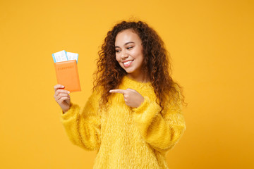 Pretty young african american girl in fur sweater posing isolated on yellow orange background. People lifestyle concept. Mock up copy space. Pointing index finger on passport boarding pass tickets.