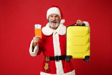 Excited elderly gray-haired mustache bearded Santa man in Christmas hat posing isolated on red background. New Year 2020 celebration concept. Mock up copy space. Holding passport, ticket and suitcase.