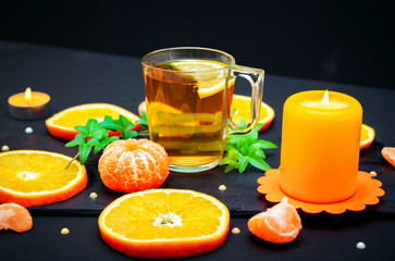 Citrus background –ripe juicy orange and tangerine slices, cup of green tea, burning candle and ivy branch with green leaves on black background.