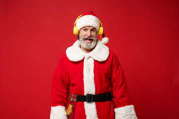 Fototapeta na wymiar Elderly gray-haired mustache bearded Santa man in Christmas hat posing isolated on red background. Happy New Year 2020 celebration holiday concept. Mock up copy space. Listening music with headphones.