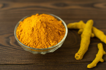 Blurred turmeric powder in glass bowl and turmeric roots on wooden background.