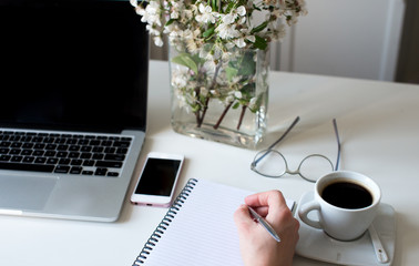 Home office desk in white colors with woman hand writing on a notebook with a pen with laptop, cup of coffee, notebook, phone, glasses on a white background. Business womans workplace and objects.