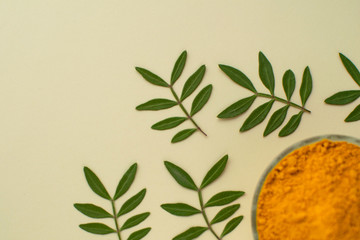 Turmeric powder in glass bowl at pastel ivory color background with green leaves. Minimalist natural backdrop.