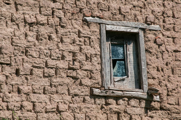 Fototapeta na wymiar Brick wall with adobe clay plaster and broken wooden window frame remained of ruined old rural country house on cloudy blue sky background