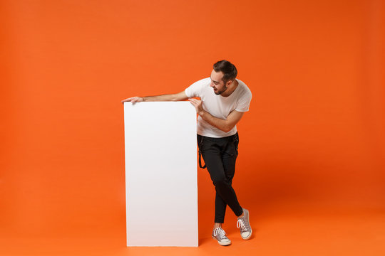 Cheerful young man in casual white t-shirt posing isolated on orange wall background. People lifestyle concept. Mock up copy space. Hold big white empty blank billboard with place for text or image.