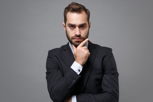 Serious young business man in classic black suit shirt tie posing isolated on grey background studio portrait. Achievement career wealth business concept. Mock up copy space. Put hand prop up on chin.