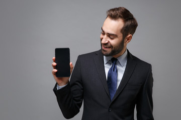 Cheerful young business man in classic suit shirt tie posing isolated on grey background. Achievement career wealth business concept. Mock up copy space. Holding mobile phone with blank empty screen.