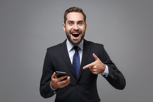 Excited Young Business Man In Classic Black Suit Shirt Tie Posing Isolated On Grey Background. Achievement Career Wealth Business Concept. Mock Up Copy Space. Pointing Index Finger On Mobile Phone.