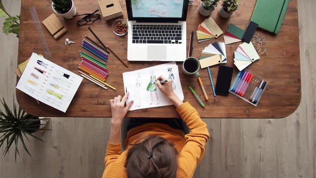Flat lay of young female clothes designer sitting at workplace at computer screen and coloring clothes sketches using colour pencils and felt-tip pens and color palettes